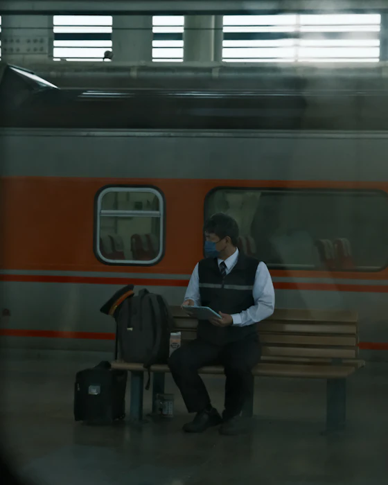 Un homme d’une quarantaine d’années attend sur un banc de gare, téléphone en main, un sac de voyage à côté
